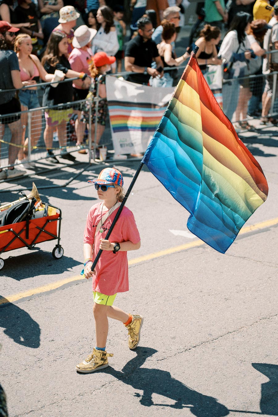A child participates in a pride parade, proudly carrying a rainbow flag while wearing a pink shirt and colorful hat.