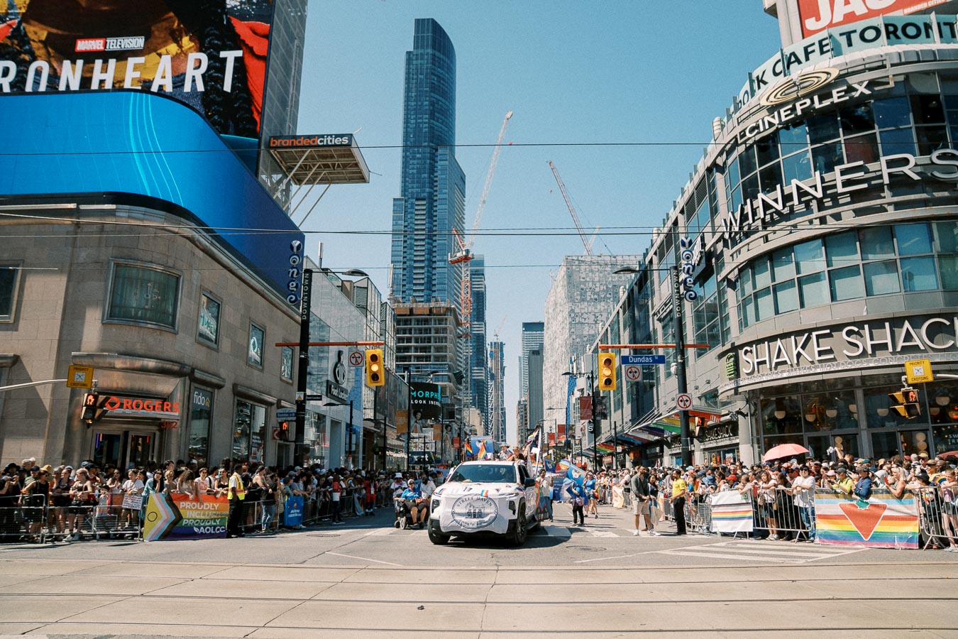 A vibrant parade takes place in downtown Toronto, with crowds lining the streets and colorful banners displayed. The scene is set against a backdrop of skyscrapers and prominent storefronts like Shake Shack and Winners.