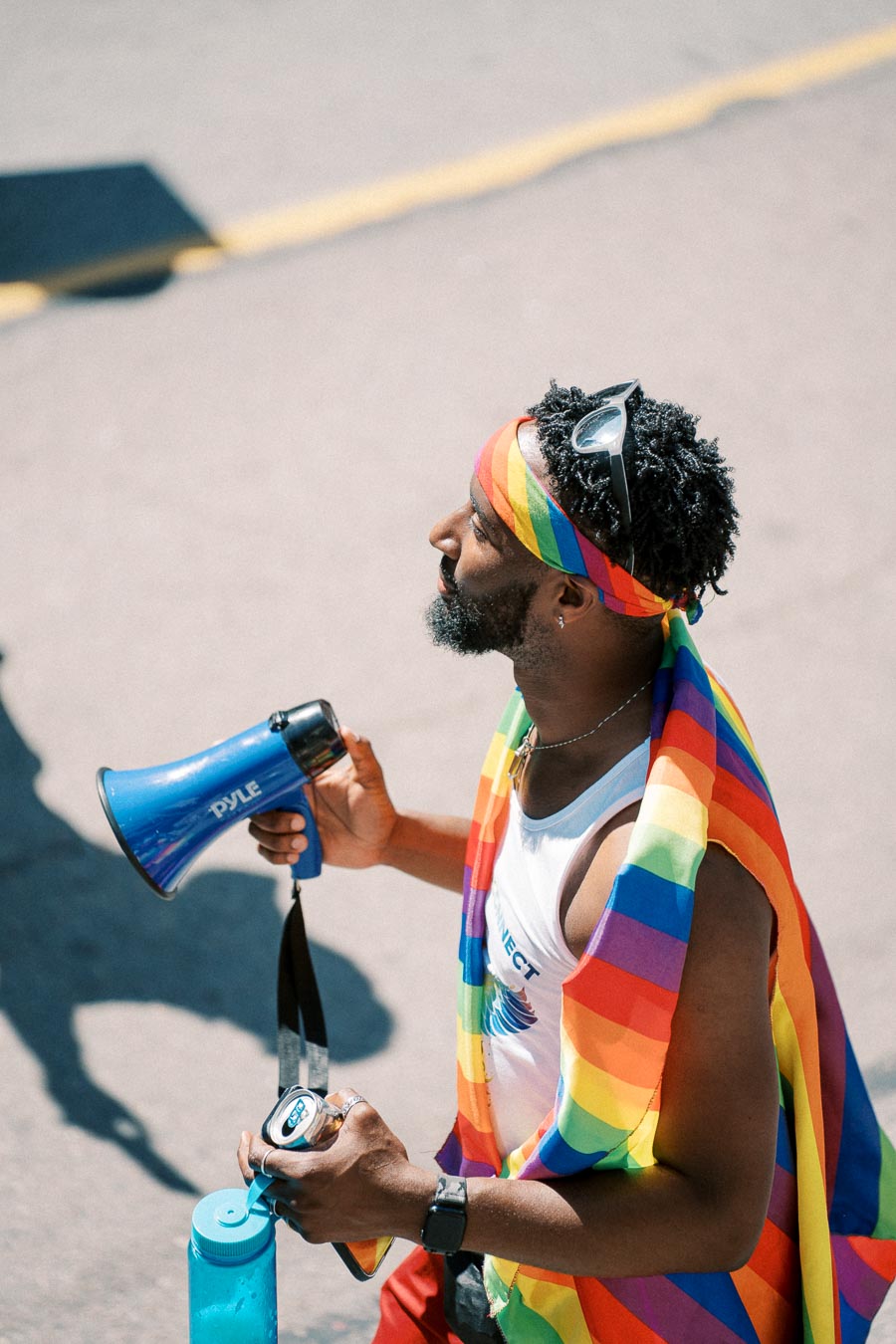 A participant at a pride event wearing a rainbow flag and headband, holding a blue megaphone and a drink.
