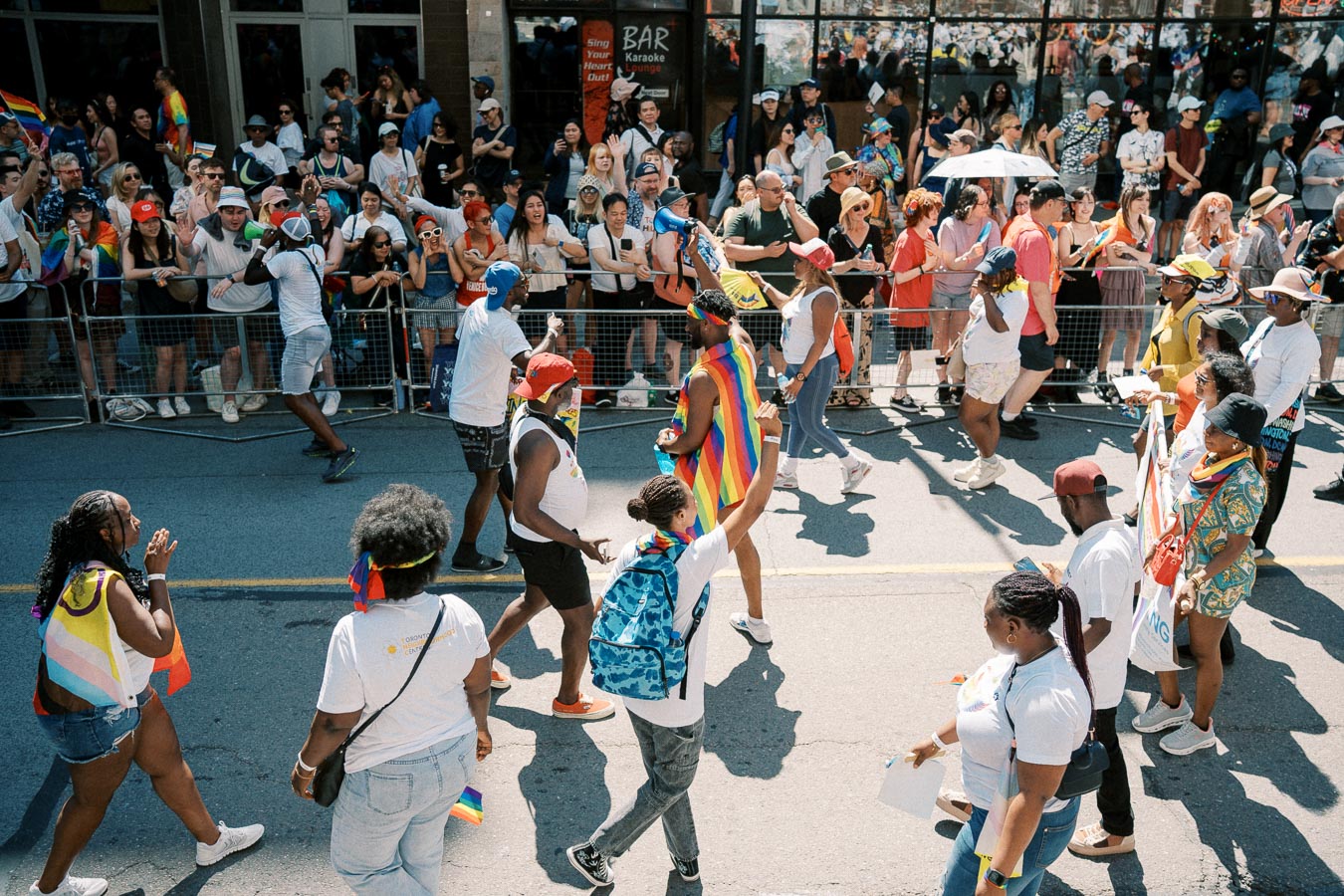 Participants in a pride parade march down the street, adorned with rainbow flags and vibrant attire, while spectators cheer from behind barriers.