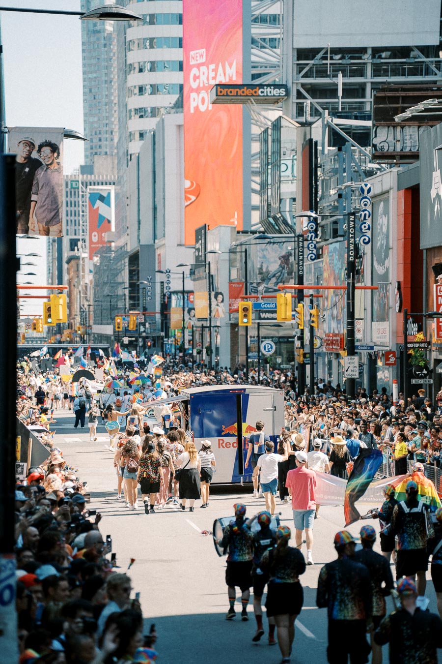 A lively parade takes place on a bustling city street, with participants carrying rainbow flags and banners, surrounded by a large crowd. The scene is set against a backdrop of tall buildings and colorful advertisements.