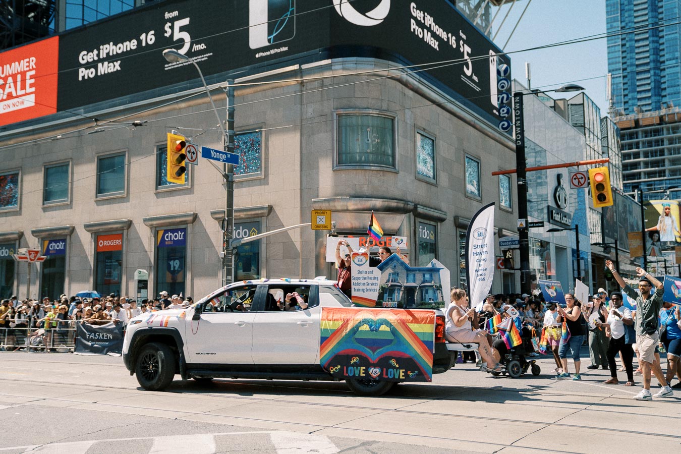 A vibrant parade scene featuring a white truck adorned with rainbow flags and participants holding signs, set against a bustling city street with a prominent iPhone advertisement.