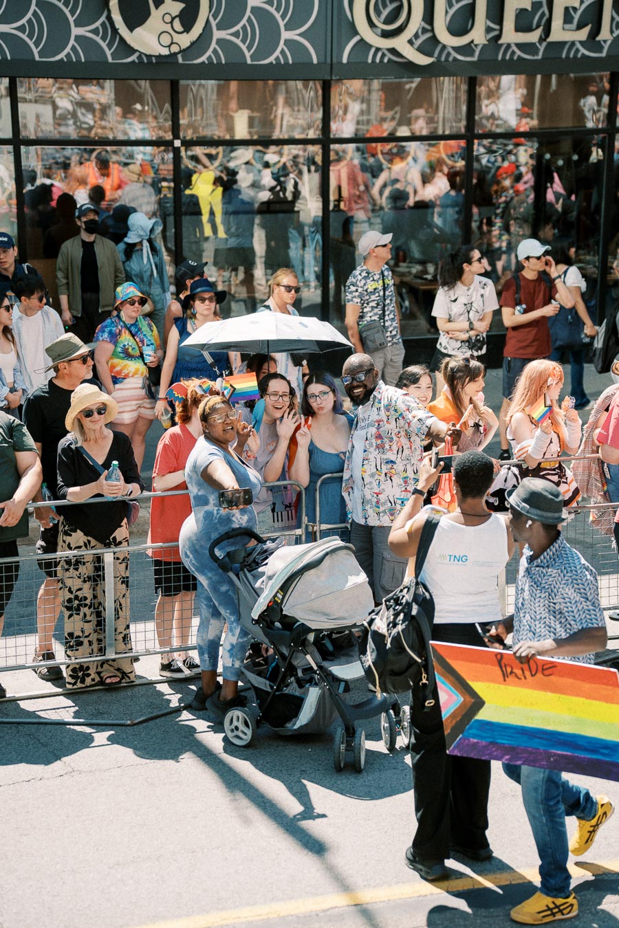 People celebrating at a pride parade, capturing moments with selfies and rainbow flags, in front of a building with reflective windows.