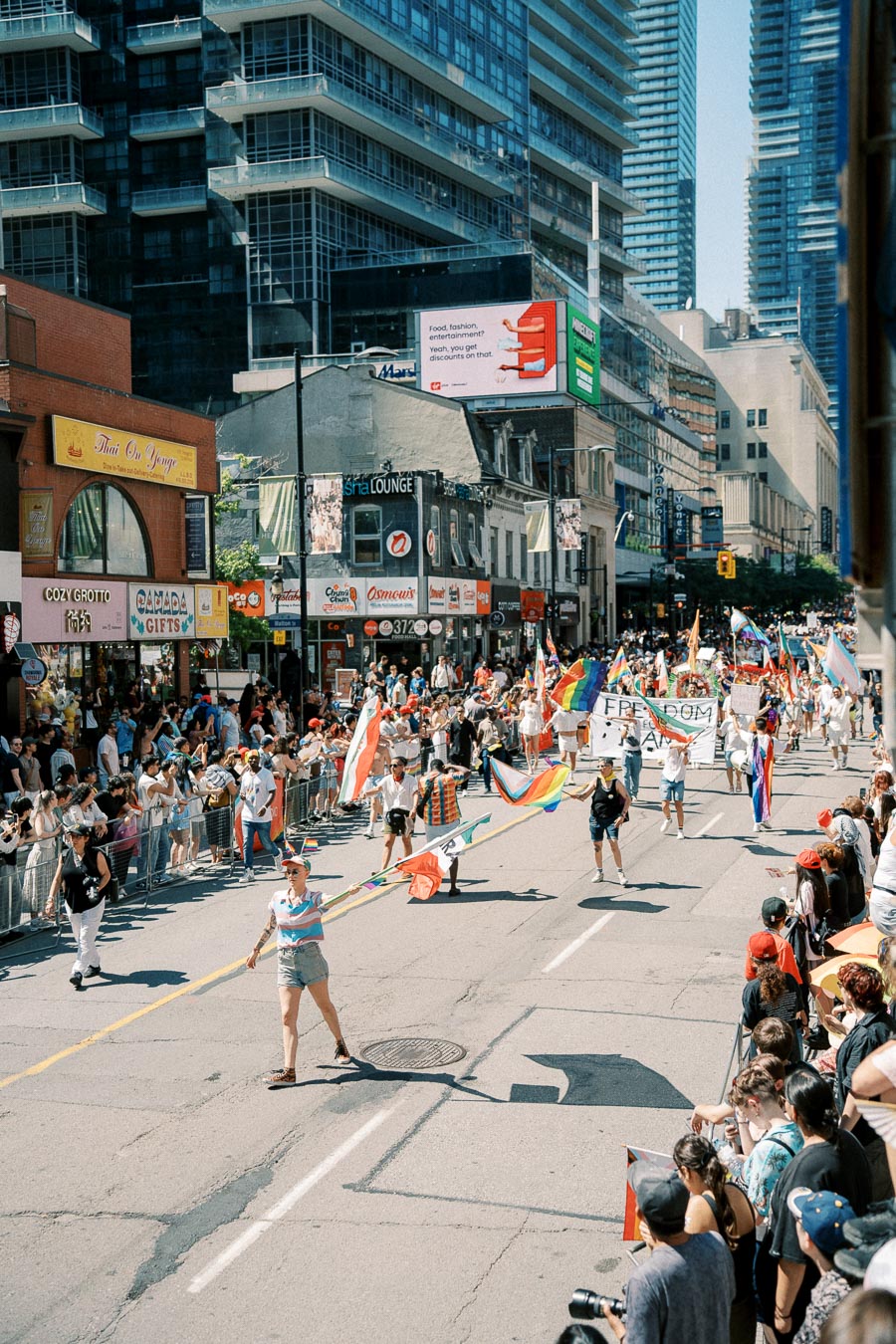 A lively parade featuring rainbow flags and banners takes place in a bustling urban street, with spectators lining the sidewalks.