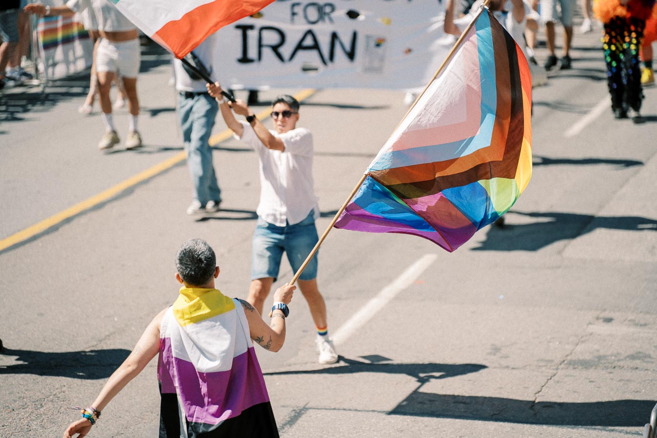 Participants in a pride parade wave a Progress Pride flag and a non-binary flag, advocating for LGBTQ+ rights.
