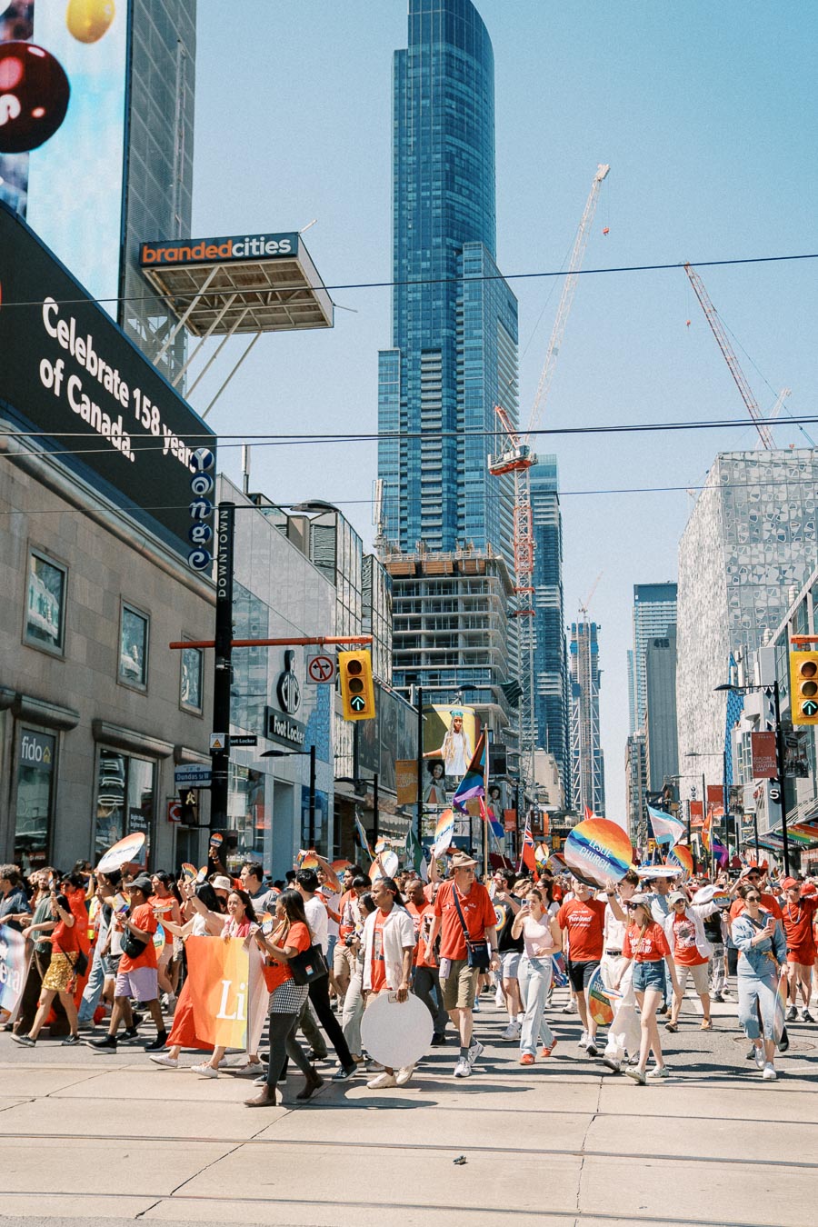 A lively parade celebrating diversity and inclusion takes place in a bustling city street, with participants holding rainbow flags and banners. The backdrop features tall skyscrapers and construction cranes, indicating urban development.