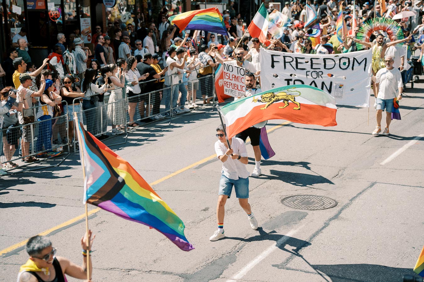Participants in a parade hold flags and banners advocating for freedom in Iran, with rainbow flags symbolizing LGBTQ+ pride.