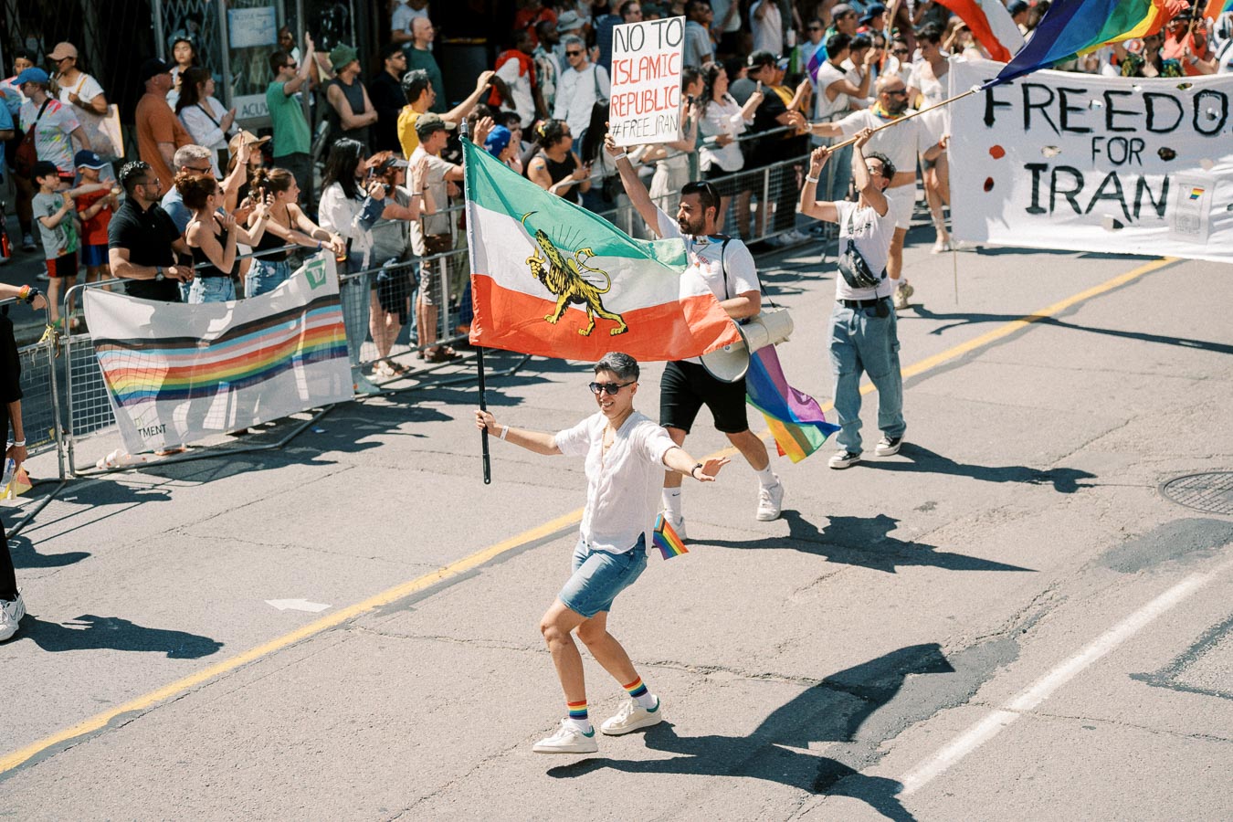 Participants in a parade hold a flag and signs advocating for freedom in Iran, with a crowd watching from the sidelines.