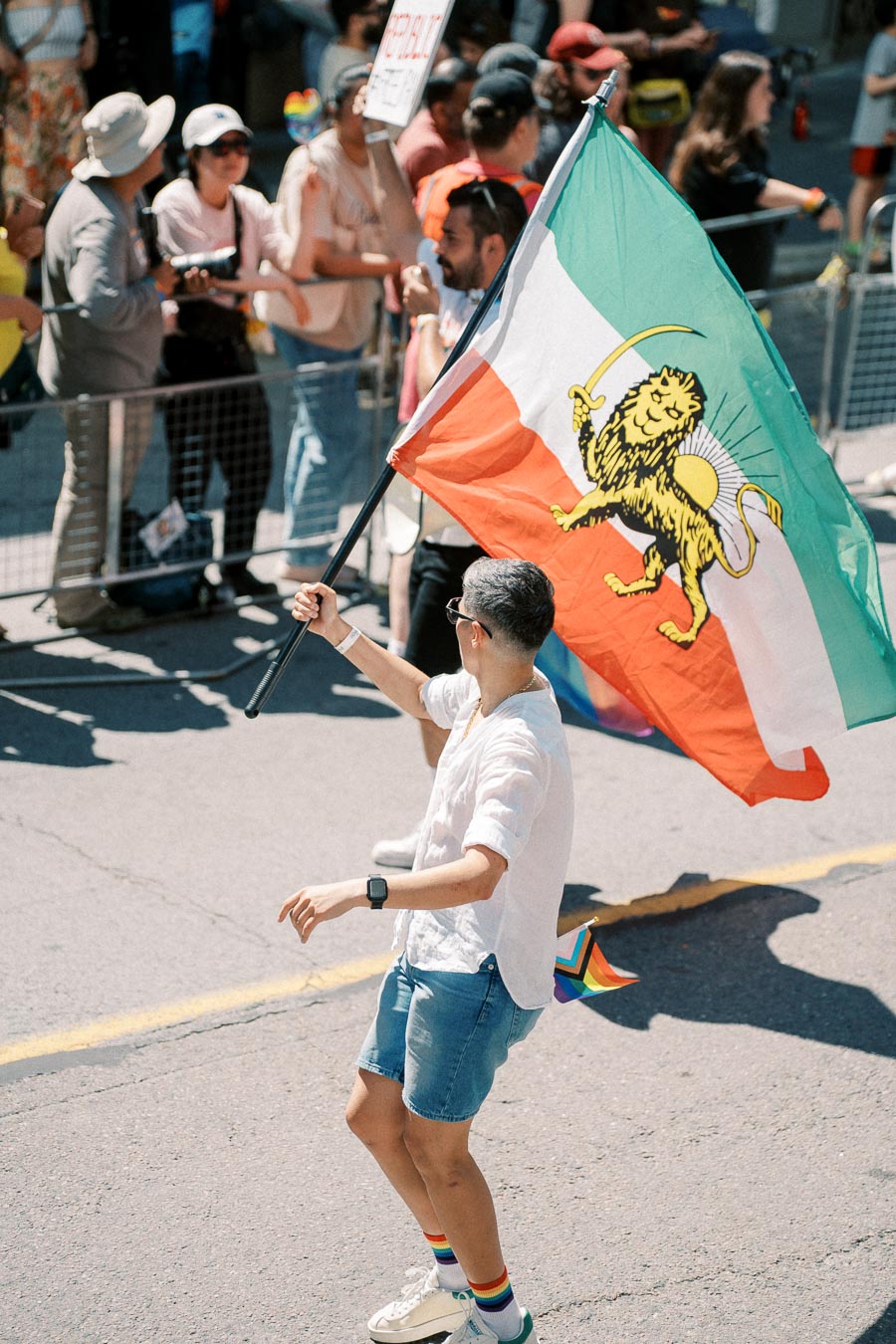 A person participates in a parade, holding a flag featuring a lion and sun emblem. The crowd watches from behind a barrier, and the participant wears casual attire with rainbow socks, suggesting a pride event.