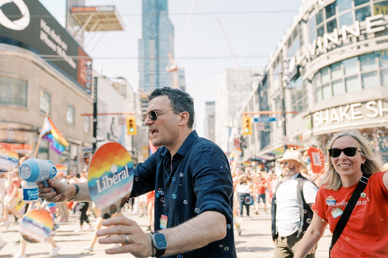 Participants in a pride parade celebrate with rainbow flags and signs, surrounded by a lively urban backdrop.