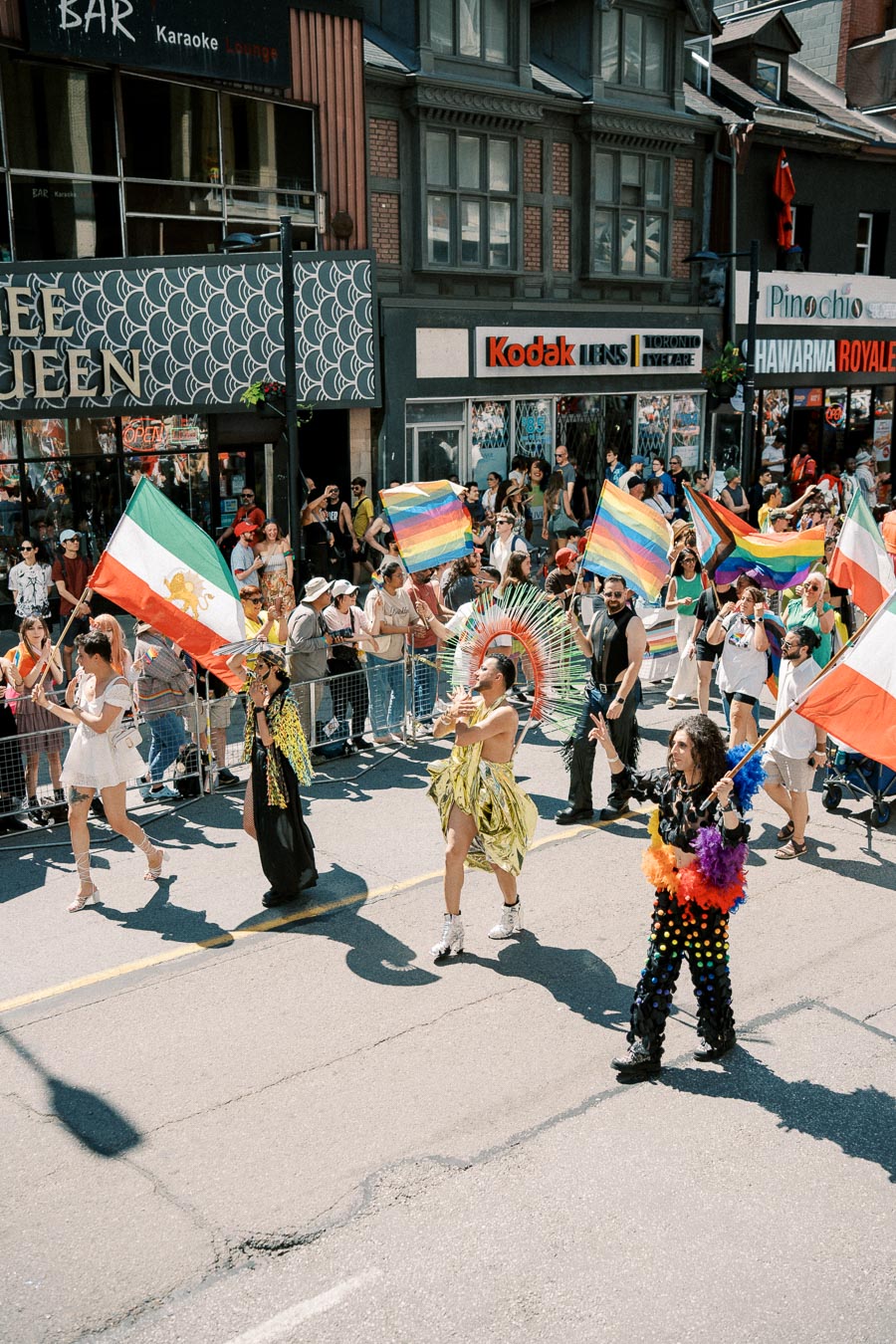 Participants in colorful costumes and holding rainbow flags march in a lively parade, with spectators lining the city street.