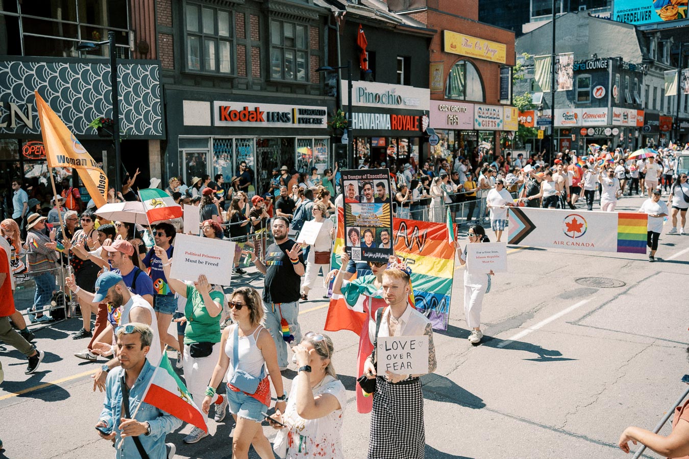 Participants in a pride parade march with signs advocating for freedom and equality, surrounded by a lively crowd and colorful flags.