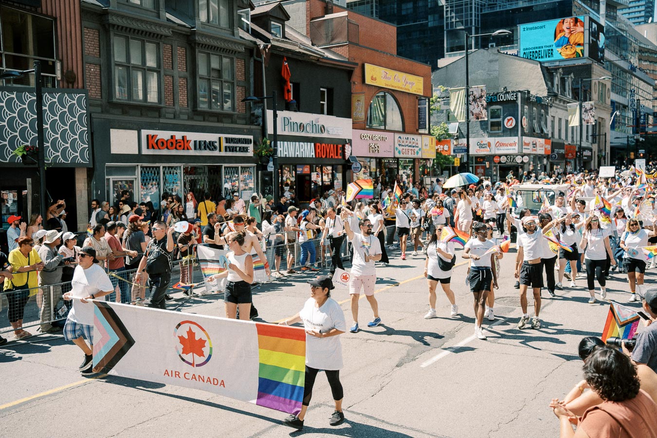 Participants in a pride parade march down a city street, holding rainbow flags and an Air Canada banner, while spectators watch from the sidewalk.
