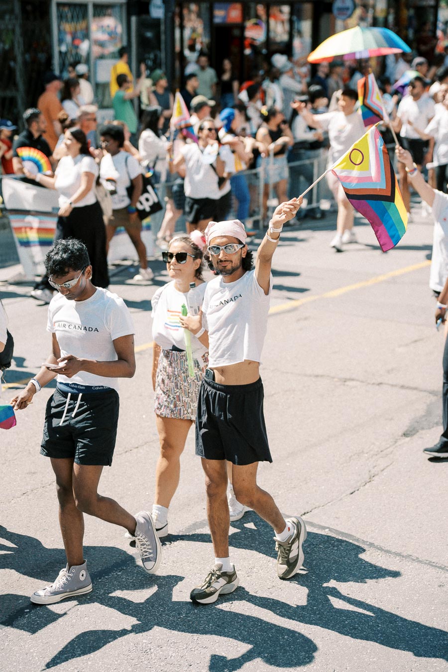 Participants in a pride parade walk down the street, one holding a rainbow flag with an intersex symbol. The crowd in the background is celebrating with colorful attire and accessories.