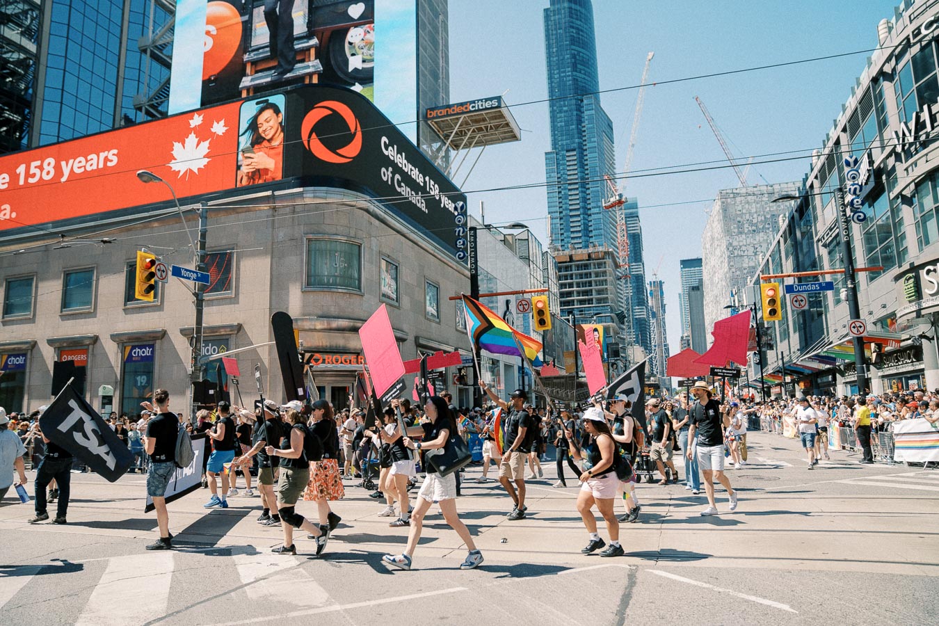 A lively parade in Toronto featuring participants with colorful flags and banners, set against the backdrop of the city's skyscrapers and digital billboards.