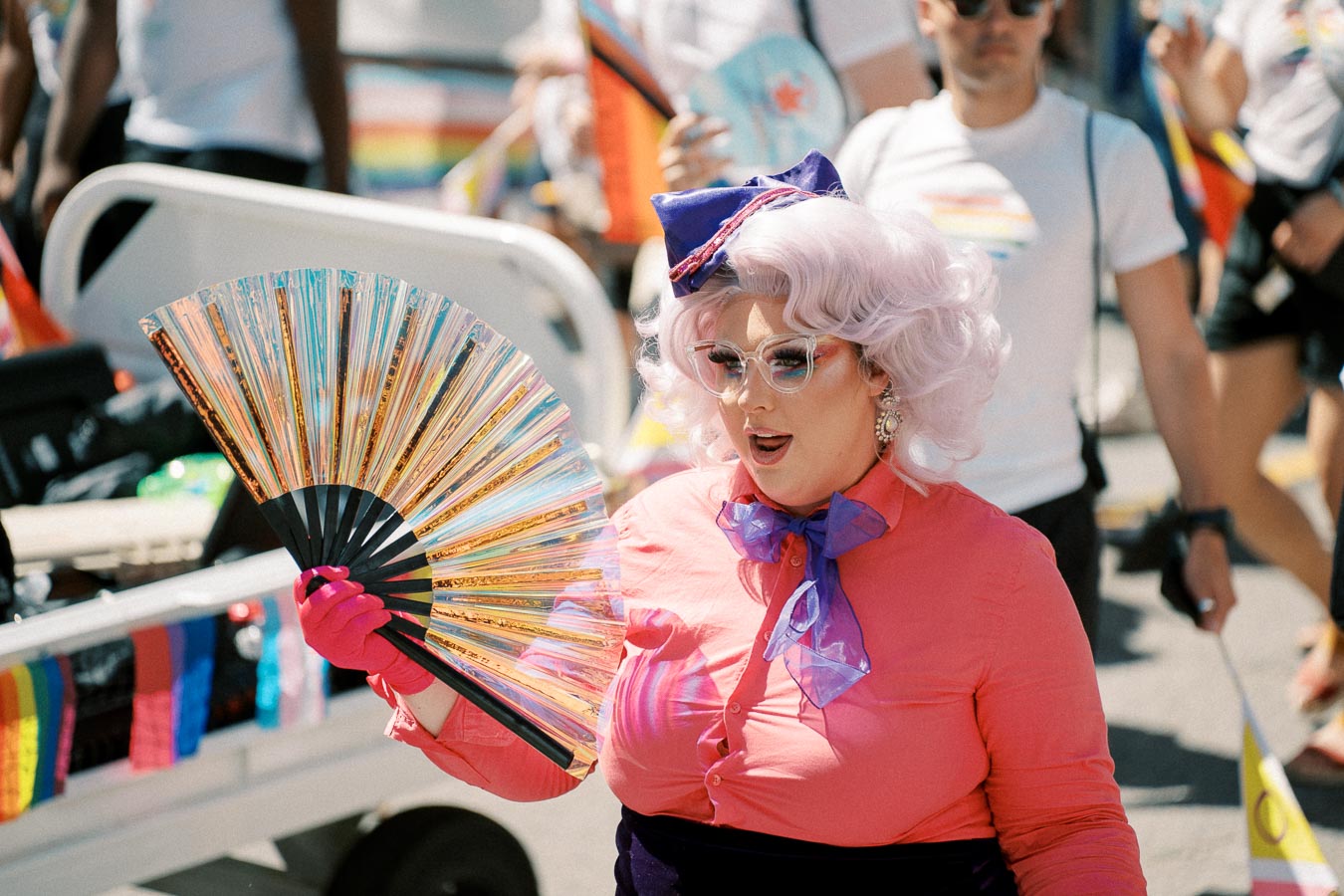 A person dressed in a vibrant pink outfit with a purple bow and white wig participates in a pride parade, holding a colorful fan. The background features people wearing white shirts with rainbow designs, indicating a festive and inclusive atmosphere.