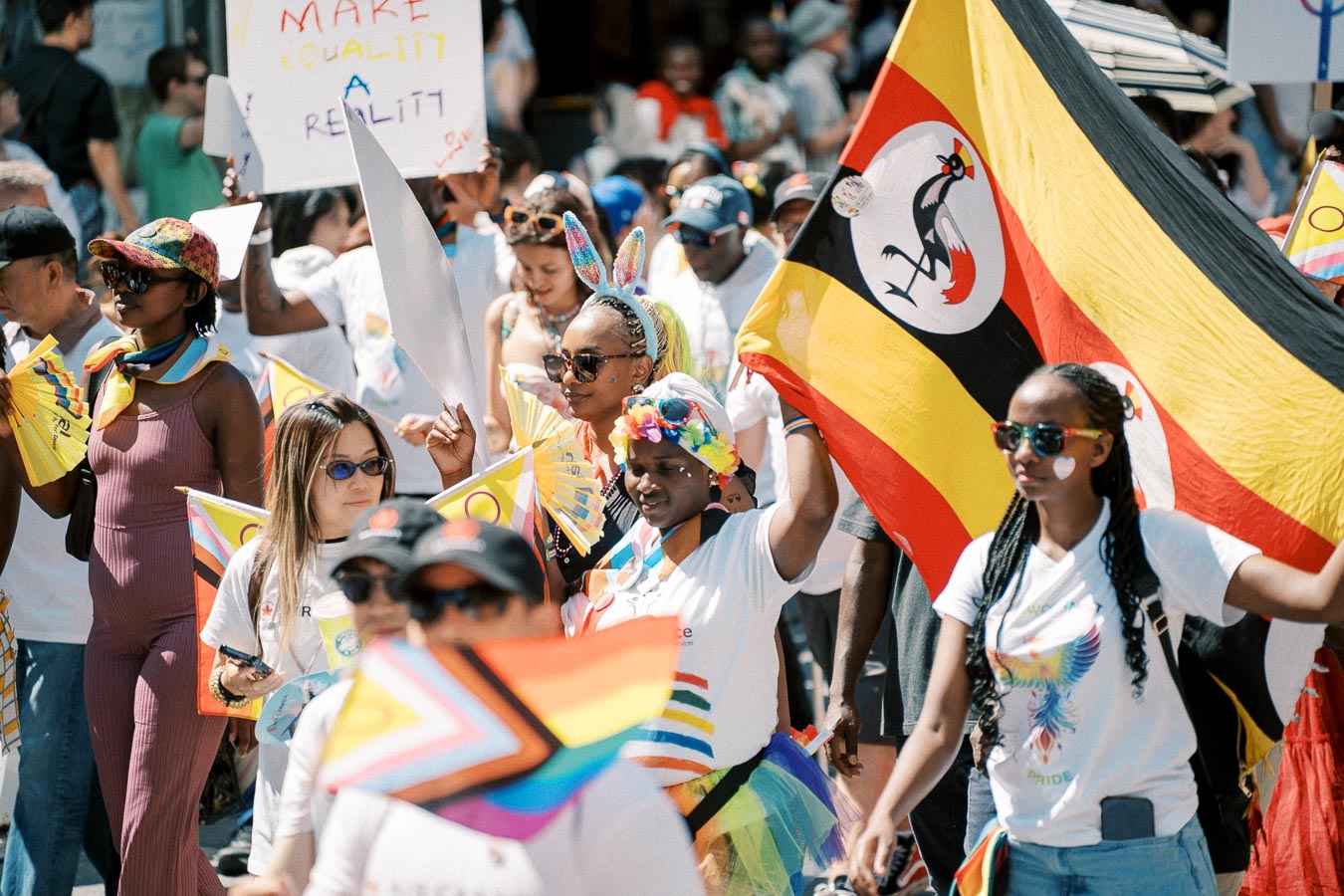 Participants in a pride parade hold flags and signs promoting equality, including a Ugandan flag, showcasing diversity and support for LGBTQ+ rights.