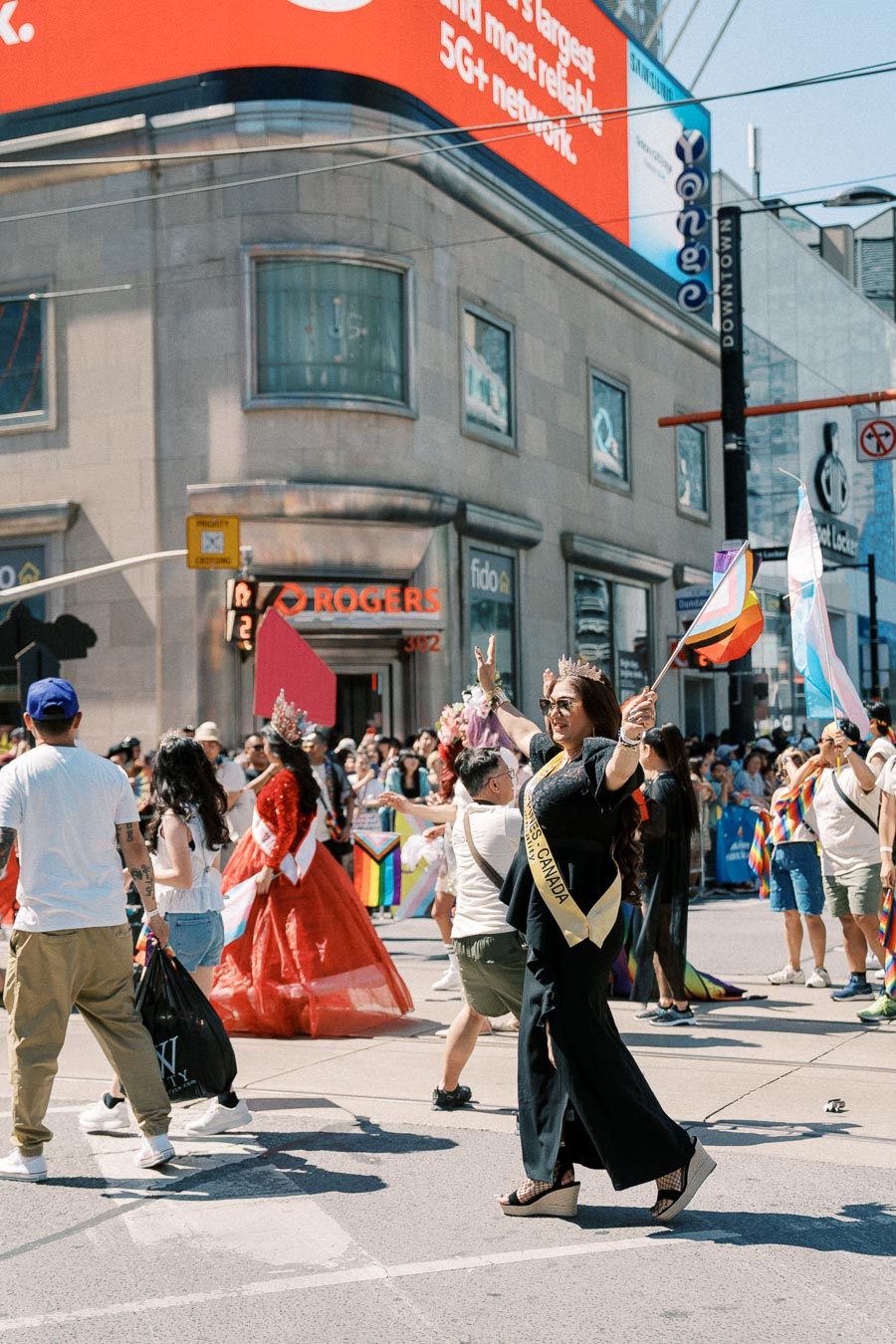 Participants celebrate at a vibrant parade, with a person wearing a 'Miss Canada' sash and others holding pride flags.