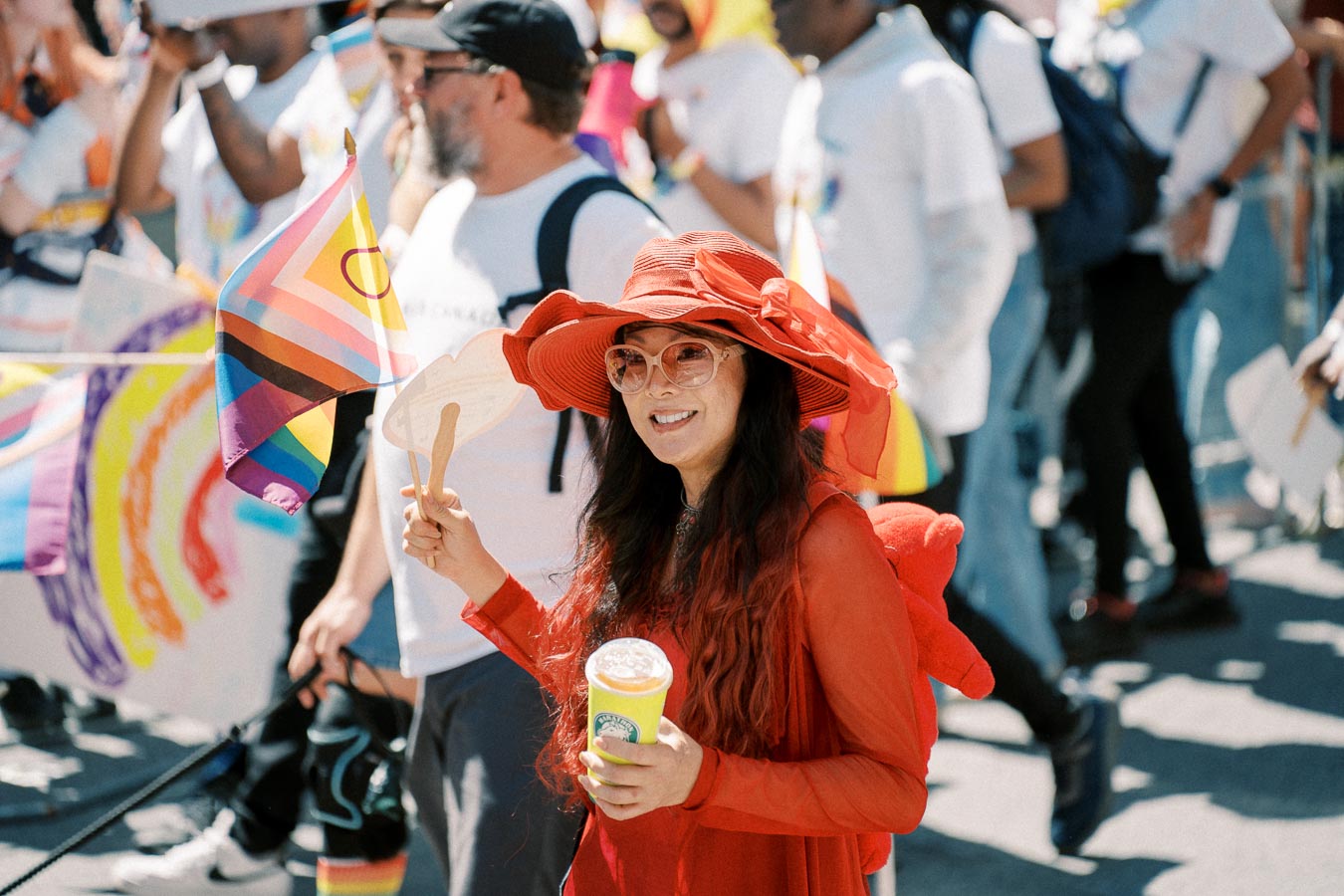 A vibrant scene at a pride parade featuring a person in a striking red outfit and hat, holding a drink and a fan, surrounded by participants waving pride flags.