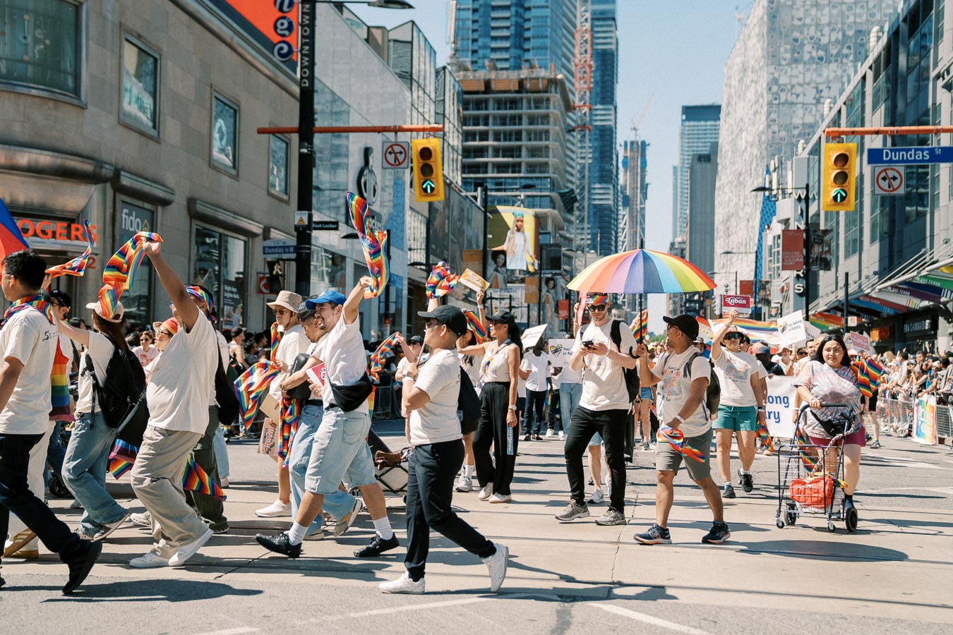 Participants celebrate at a pride parade, waving rainbow flags and carrying colorful umbrellas on a bustling city street.