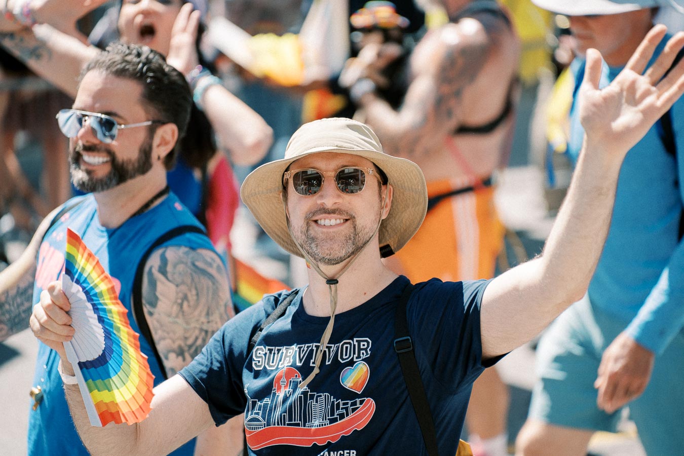 A cheerful participant at a pride parade, wearing a sun hat and sunglasses, waves while holding a rainbow fan. Their t-shirt features a rainbow heart and the word 'Survivor'.