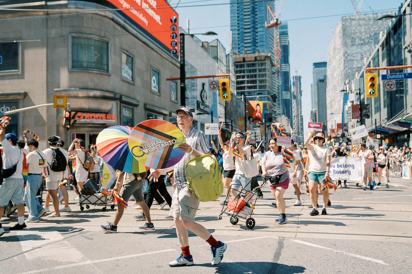 Participants in a pride parade march through a city street, displaying rainbow flags and signs. The event takes place in an urban setting with visible buildings and a Rogers store.