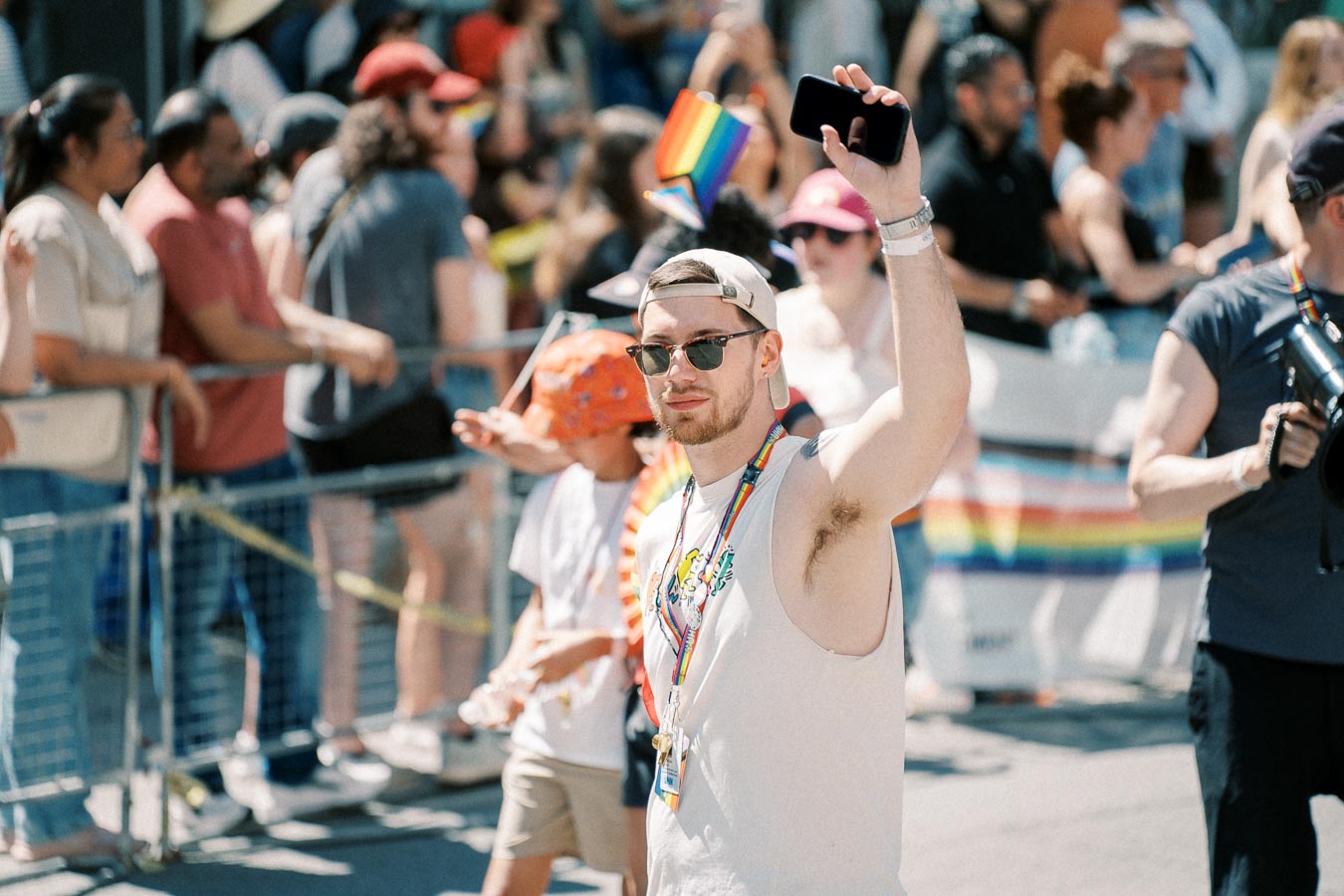 A participant at a pride parade raises a phone in celebration, surrounded by a crowd and rainbow flags.