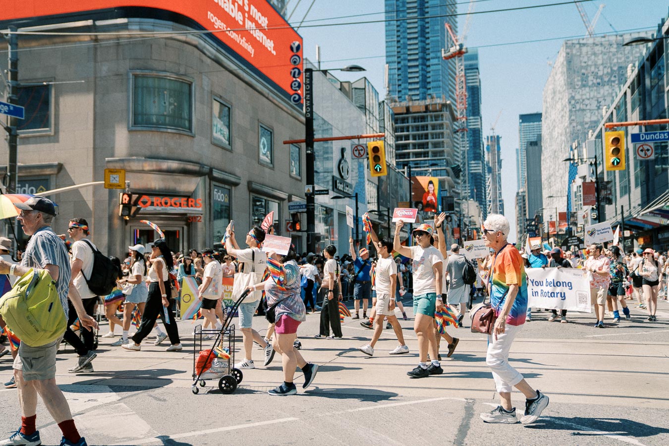 Participants in a pride parade march through a bustling city street, waving rainbow flags and holding signs, with skyscrapers and a prominent Rogers sign in the background.