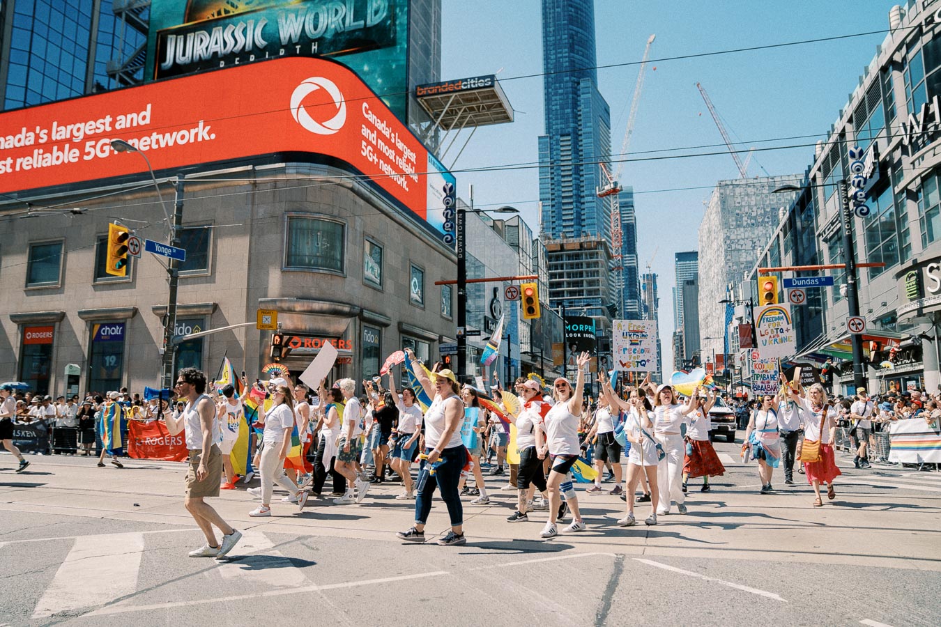 A lively parade in downtown Toronto, featuring participants celebrating diversity and freedom with signs and flags. The event takes place at the intersection of Yonge and Dundas Streets, with a backdrop of skyscrapers and digital billboards.