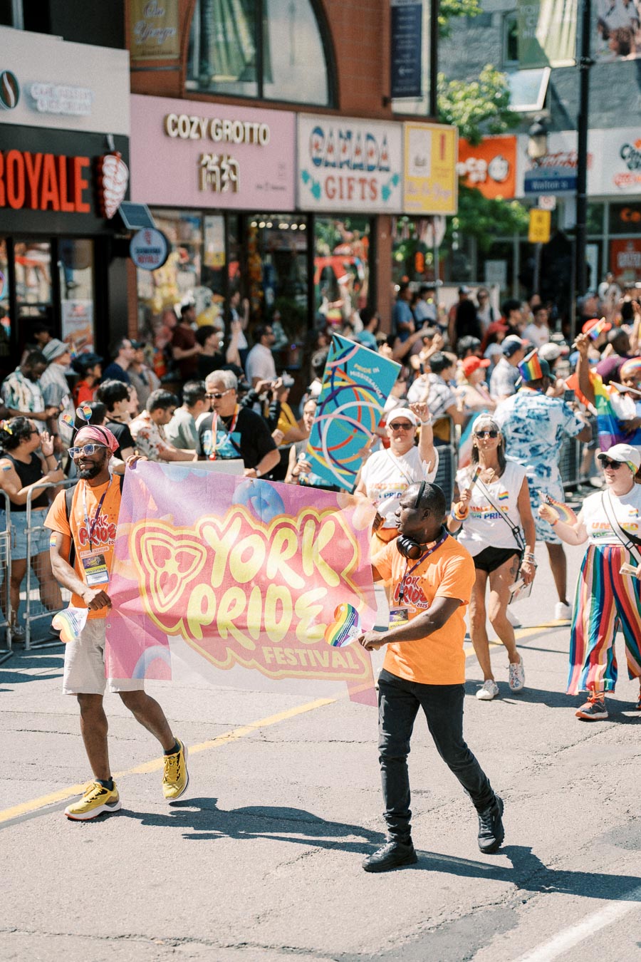 Participants in the York Pride Festival parade hold a vibrant banner as they march down a street lined with spectators and shops.