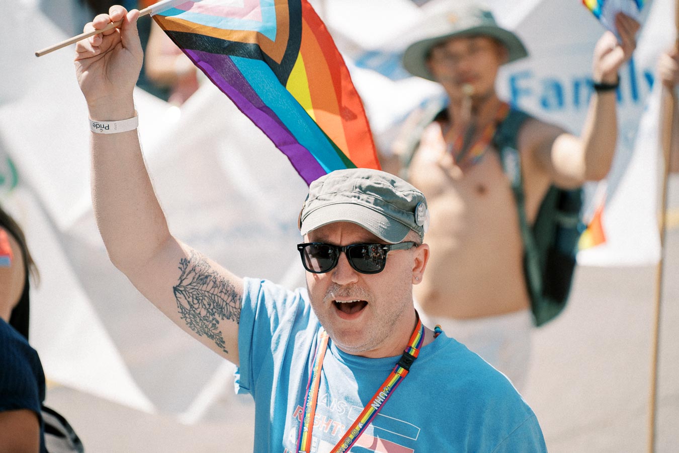 A participant at a pride parade holds a rainbow flag, wearing sunglasses and a cap, with a colorful lanyard around their neck.