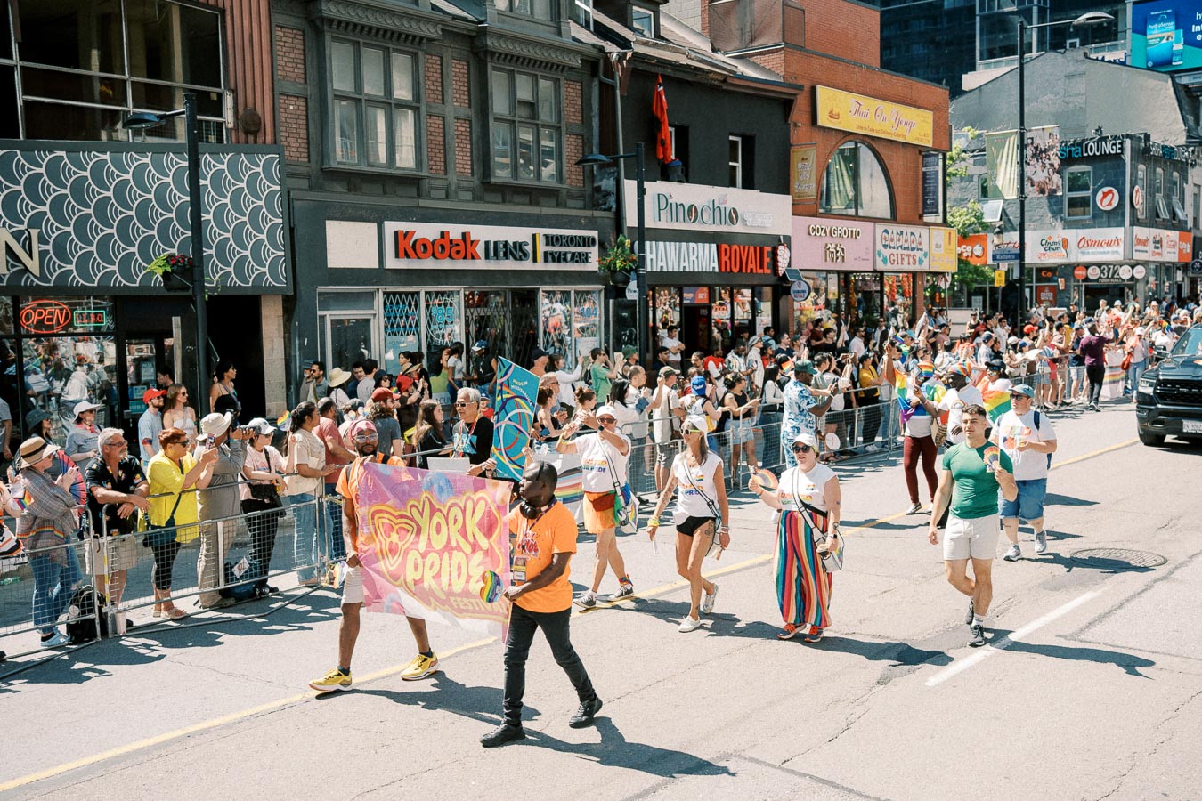 Participants march in a lively parade holding a 'York Pride Festival' banner, with a crowd of spectators lining the city street.