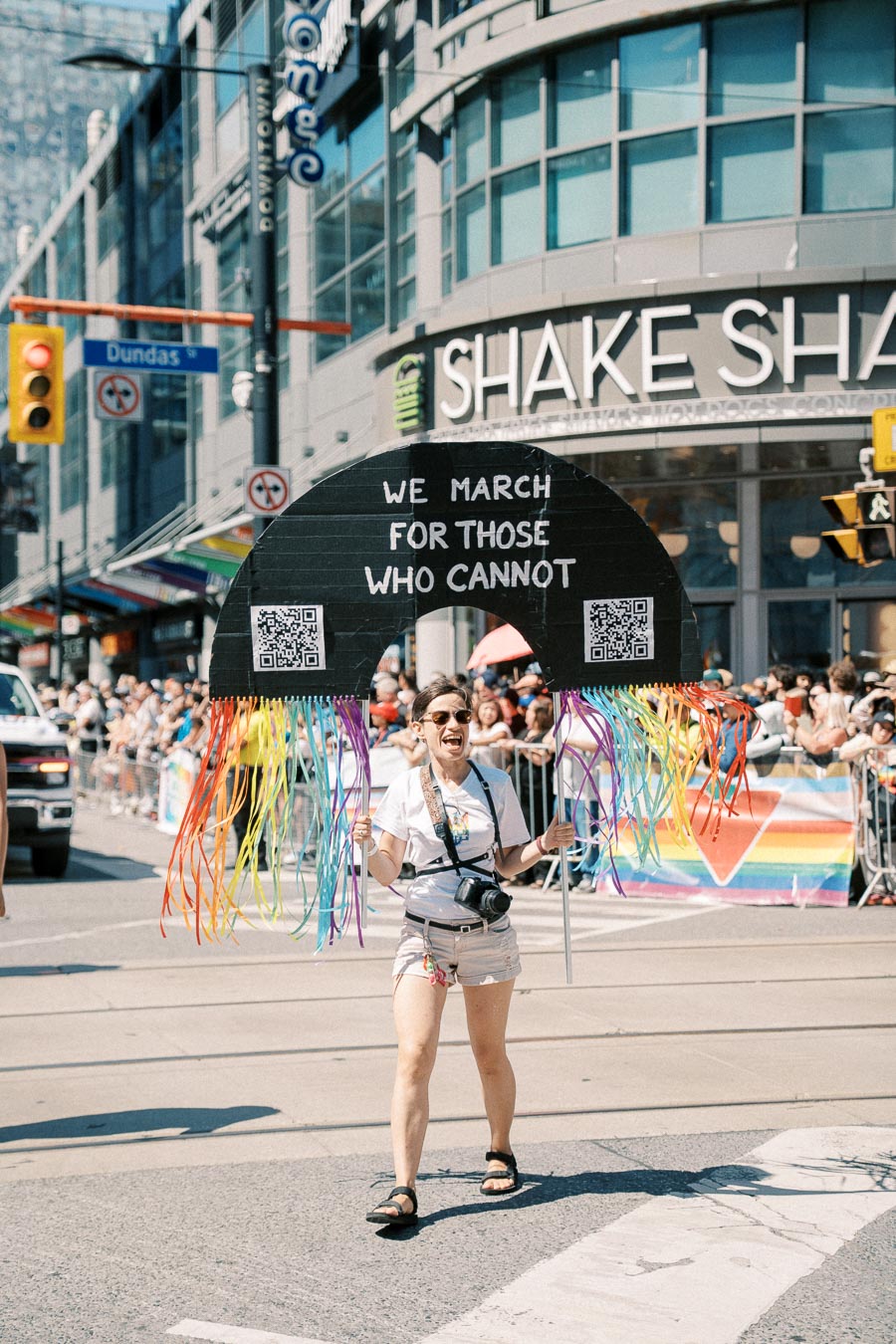 A participant at a pride parade holds a sign reading "We March For Those Who Cannot" with rainbow ribbons, in front of a Shake Shack restaurant.