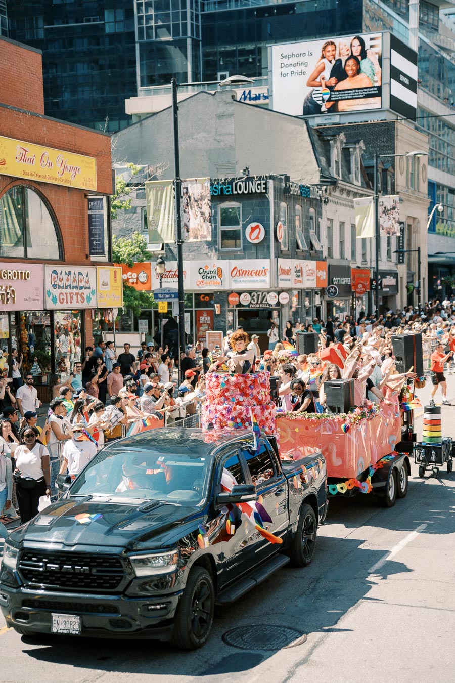 A vibrant parade float adorned with flowers and rainbow flags moves through a city street, attracting a crowd of onlookers.