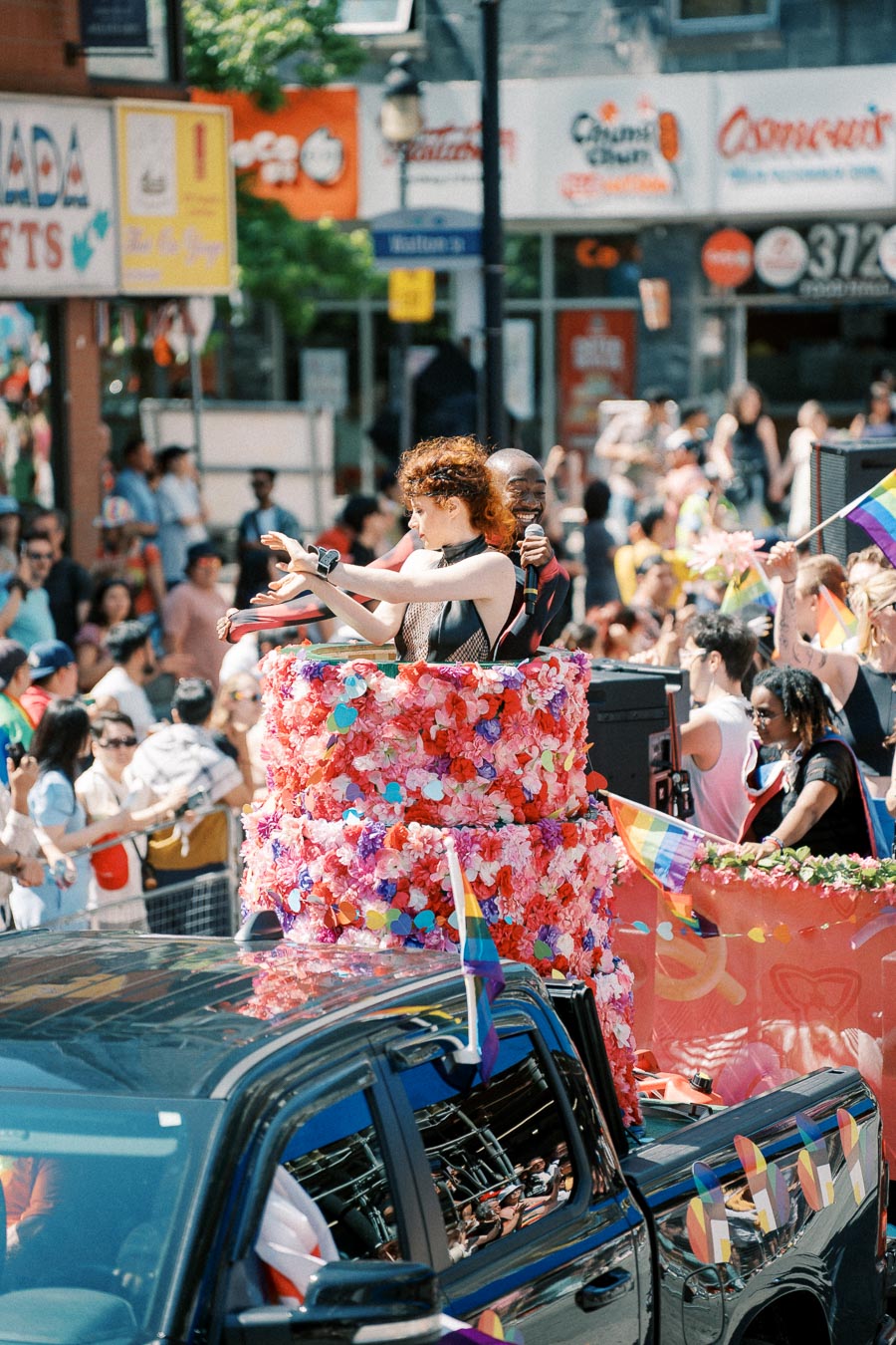 A colorful parade float adorned with flowers and rainbow flags moves through a lively crowd during a pride parade.