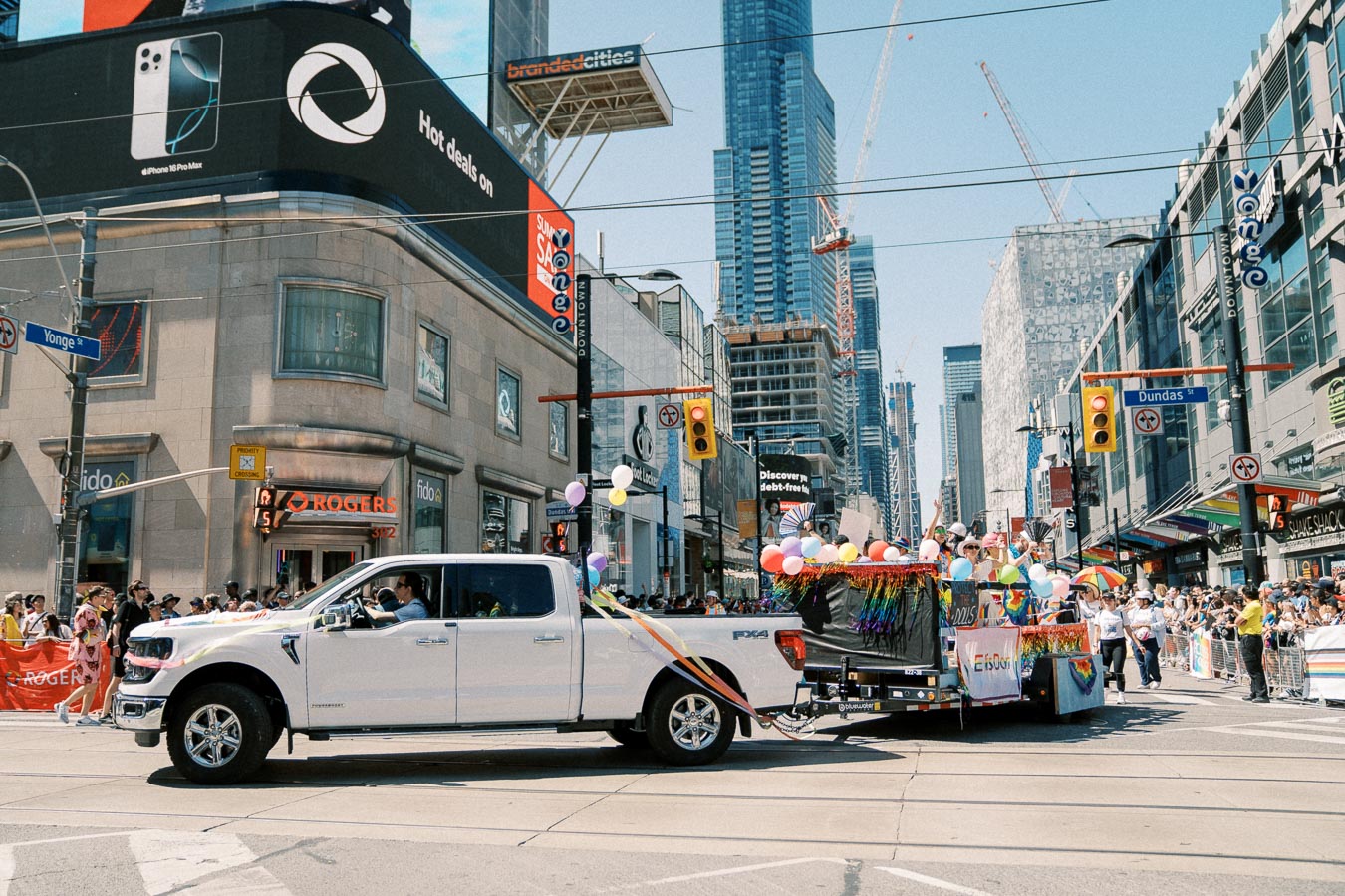 A vibrant parade scene in downtown Toronto, featuring a white pickup truck towing a colorful float adorned with balloons and rainbow decorations. The event takes place at the intersection of Yonge and Dundas Streets, with crowds lining the sidewalks and skyscrapers towering above.