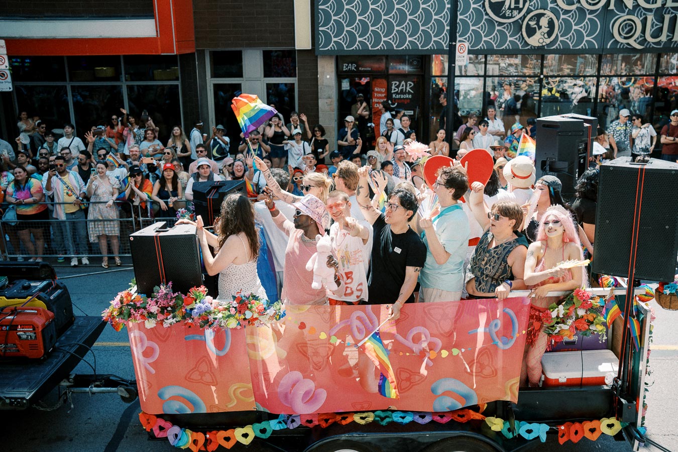 A lively scene from a parade featuring a float with people celebrating diversity and pride, waving rainbow flags and adorned in colorful attire.
