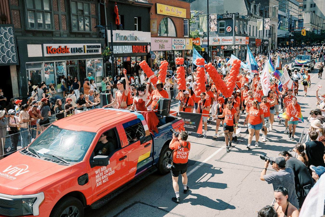 Participants in a parade wearing red shirts and carrying red balloons, promoting awareness about HIV and AIDS, as they walk alongside a red truck. Spectators watch from the sidelines.