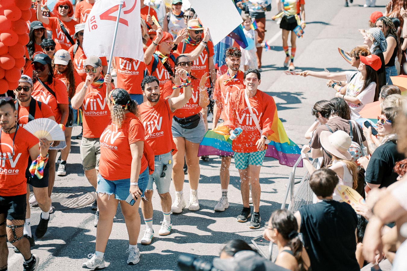 Participants in a pride parade wearing red ViiV Healthcare shirts, celebrating with rainbow flags and banners.
