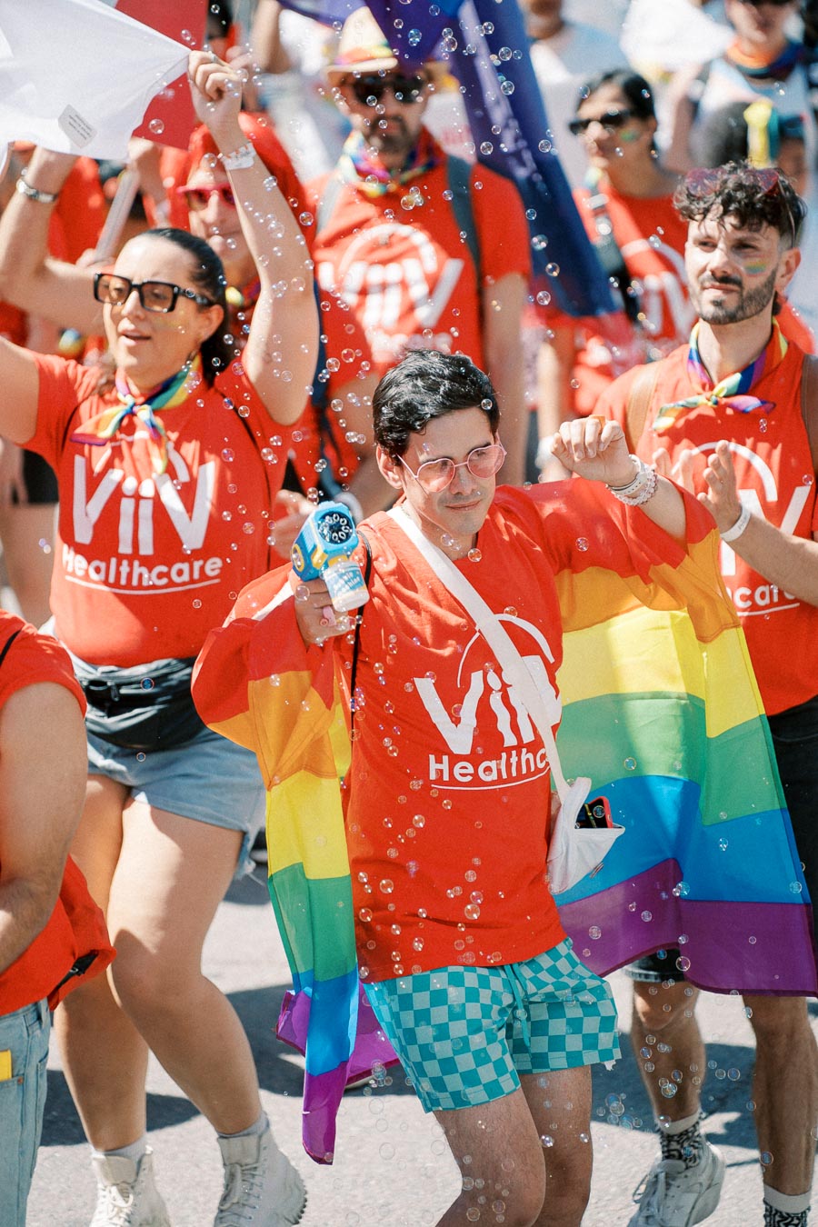 Participants in a parade wearing red ViiV Healthcare shirts, with one person holding a rainbow flag and a bubble gun, surrounded by bubbles.