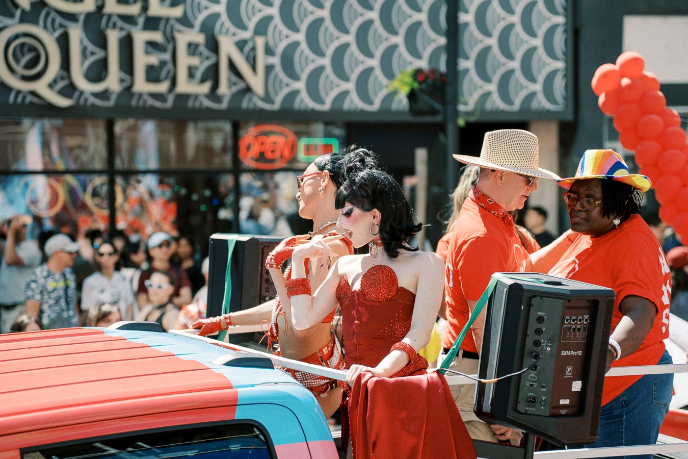 Participants in colorful costumes and hats enjoy a lively parade, with a crowd and a 'QUEEN' sign in the background.