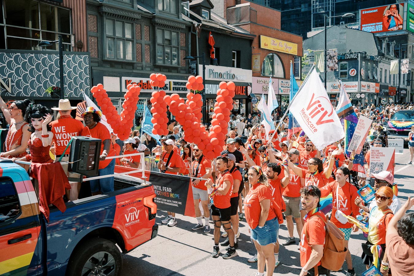 Participants in a lively parade march down a city street, wearing red shirts and holding flags and balloons spelling 'ViiV'. A colorful truck is part of the procession, and the street is lined with spectators.