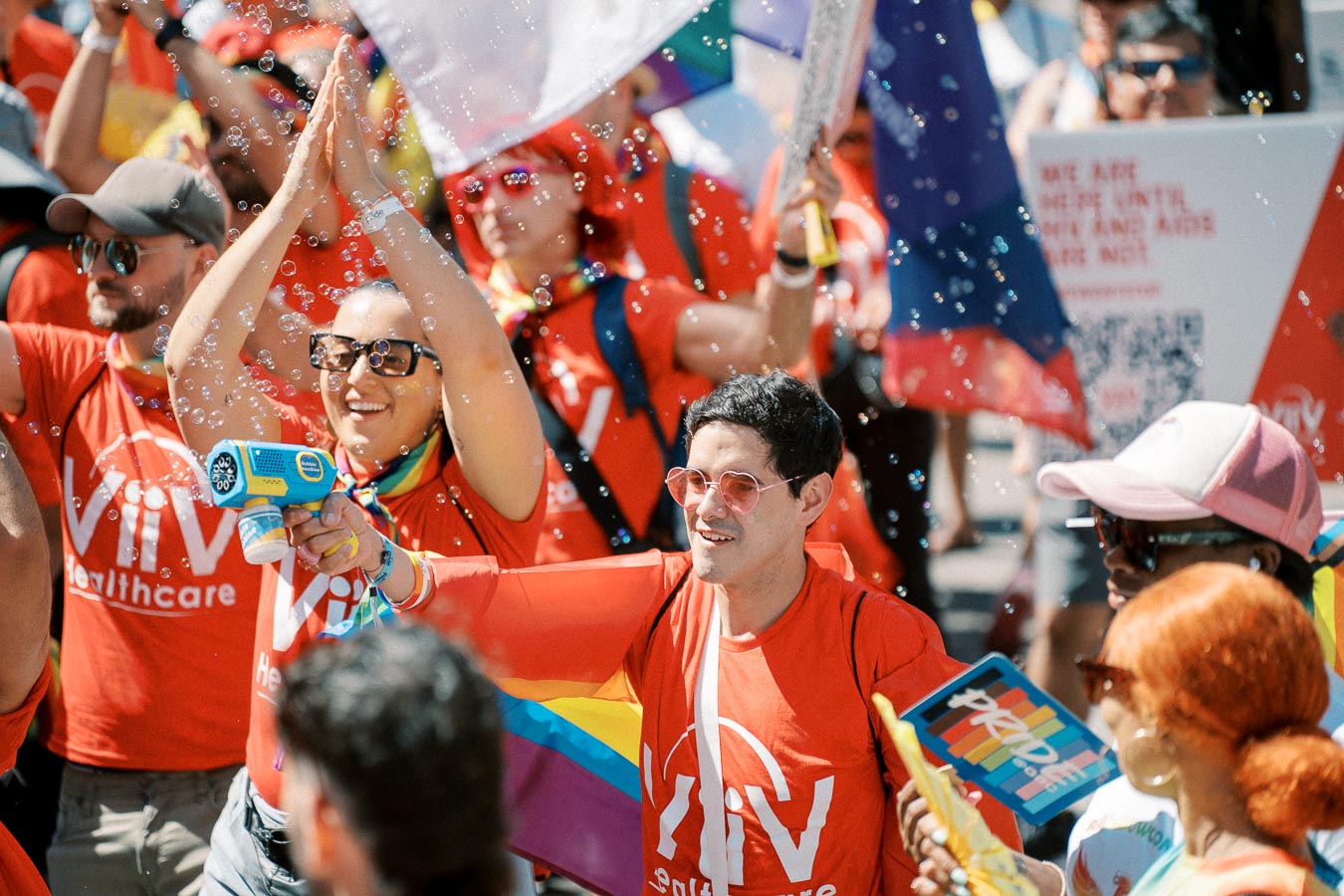 Participants in a pride parade celebrate with rainbow flags and bubbles, wearing red 'ViiV Healthcare' shirts.