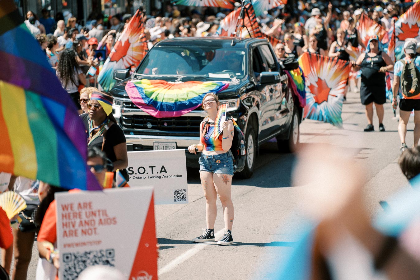 Participants celebrate at a pride parade, showcasing rainbow flags and banners. A black vehicle is adorned with a rainbow flag, and a sign from the Southern Ontario Twirling Association is visible.