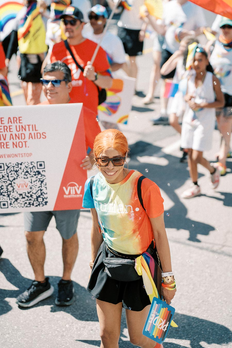 A participant in a pride parade wearing a rainbow tie-dye shirt with 'human' printed on it, smiling and holding a pride-themed fan. The parade features colorful signs and attire, celebrating diversity and inclusion.