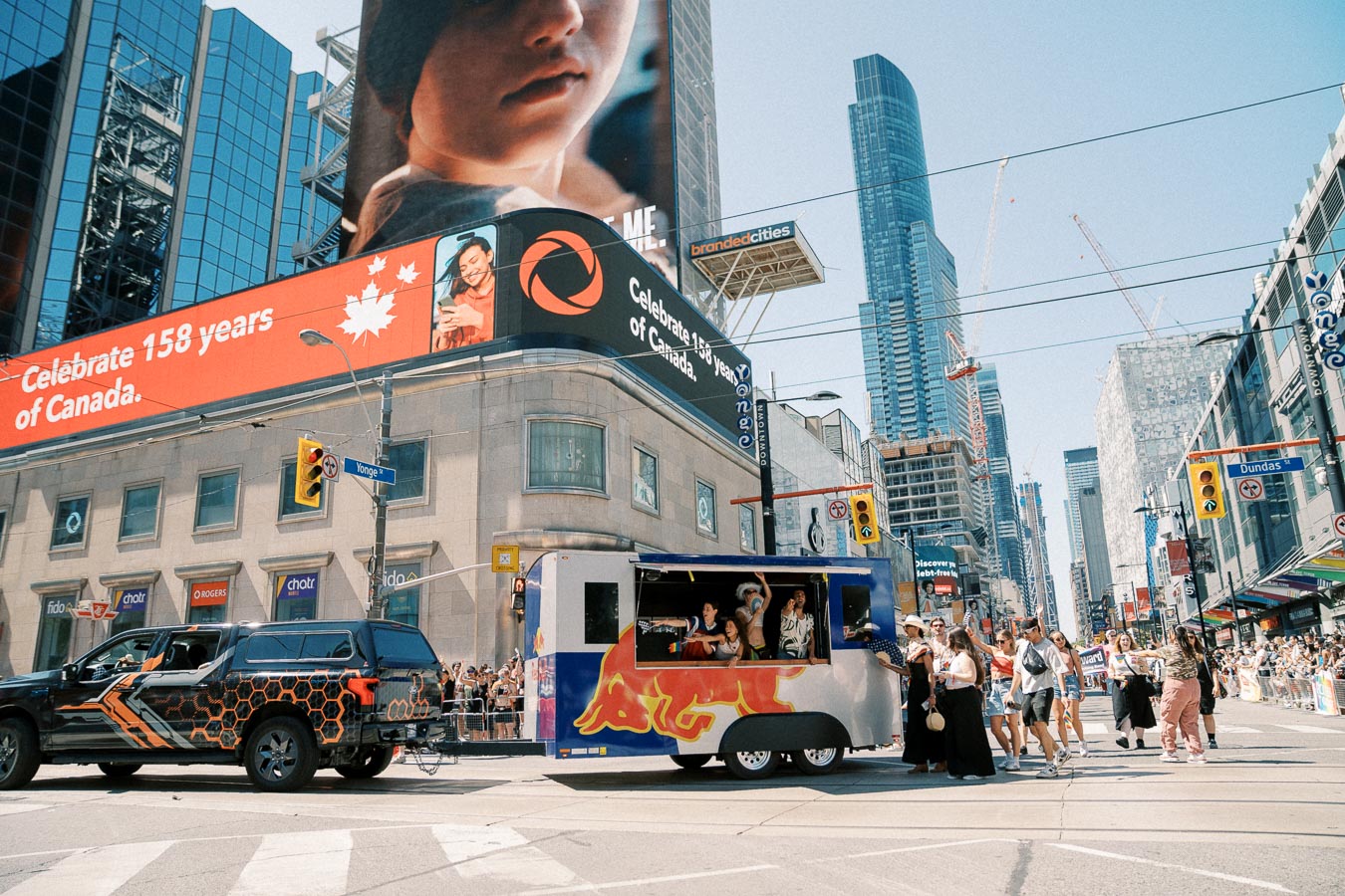 A vibrant street scene in downtown Toronto during a parade, featuring a Red Bull float and crowds celebrating. The backdrop includes skyscrapers and digital billboards.
