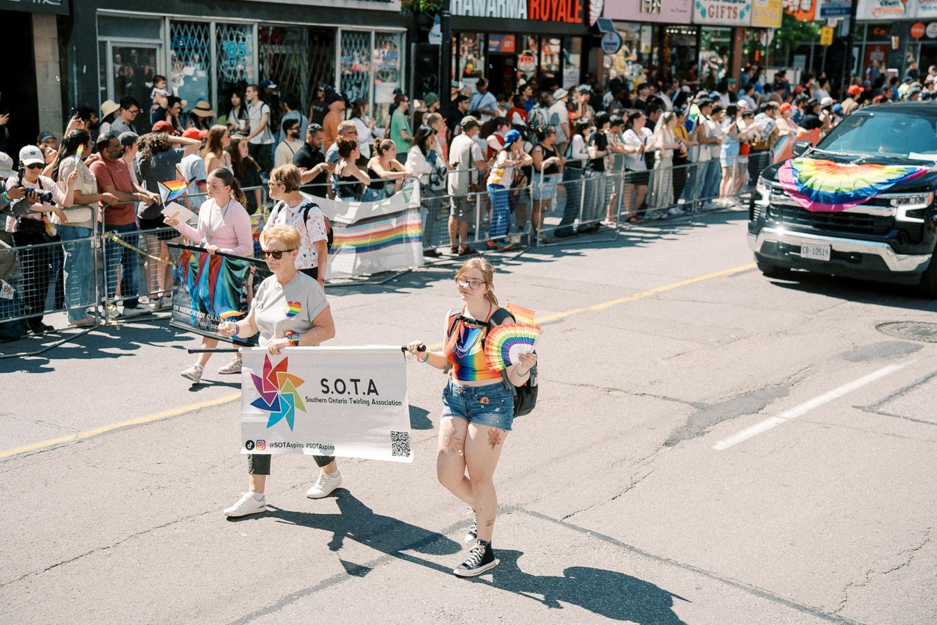 Participants from the Southern Ontario Twirling Association march in a pride parade, holding a banner and rainbow flags, with a crowd of spectators lining the street.