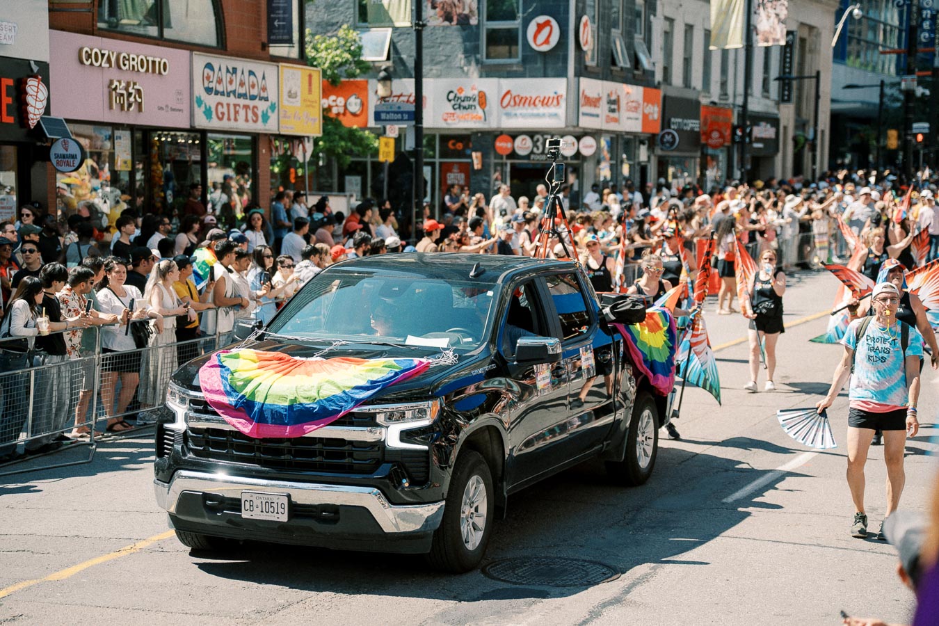 A vibrant parade scene featuring a black pickup truck adorned with rainbow flags, surrounded by participants and spectators celebrating with colorful attire and flags.