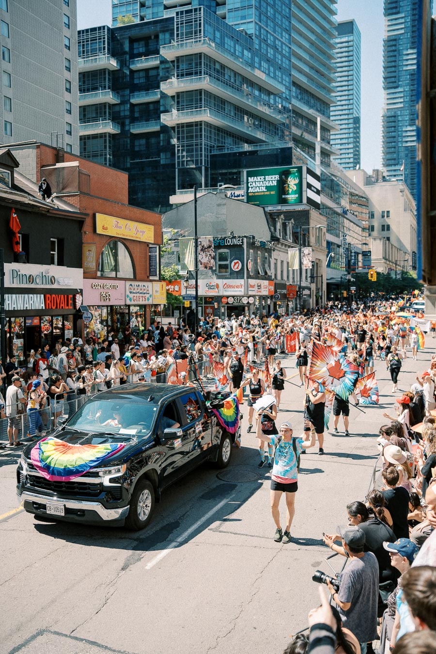 A lively parade featuring a black truck adorned with rainbow flags and participants carrying Canadian flags with rainbow designs. The event takes place on a bustling city street lined with spectators and tall buildings.