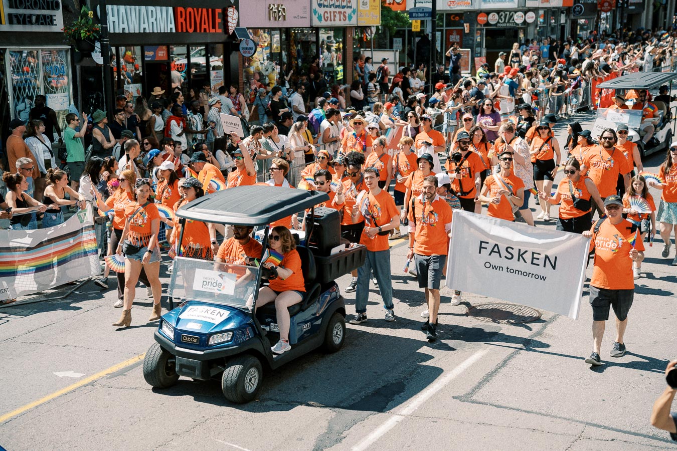 Participants in a pride parade wearing orange shirts and holding a 'FASKEN' banner walk down a crowded street, celebrating diversity and inclusion.